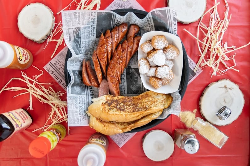 Overhead view of grilled sausage, beignets, and bread on a newspaper-lined tray with red background.